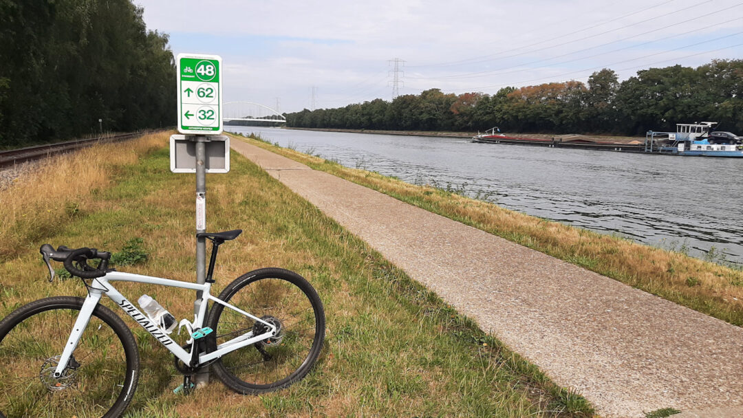 A bicycle leaning against a cycle path node sign alongside a concrete path next to a wide canal