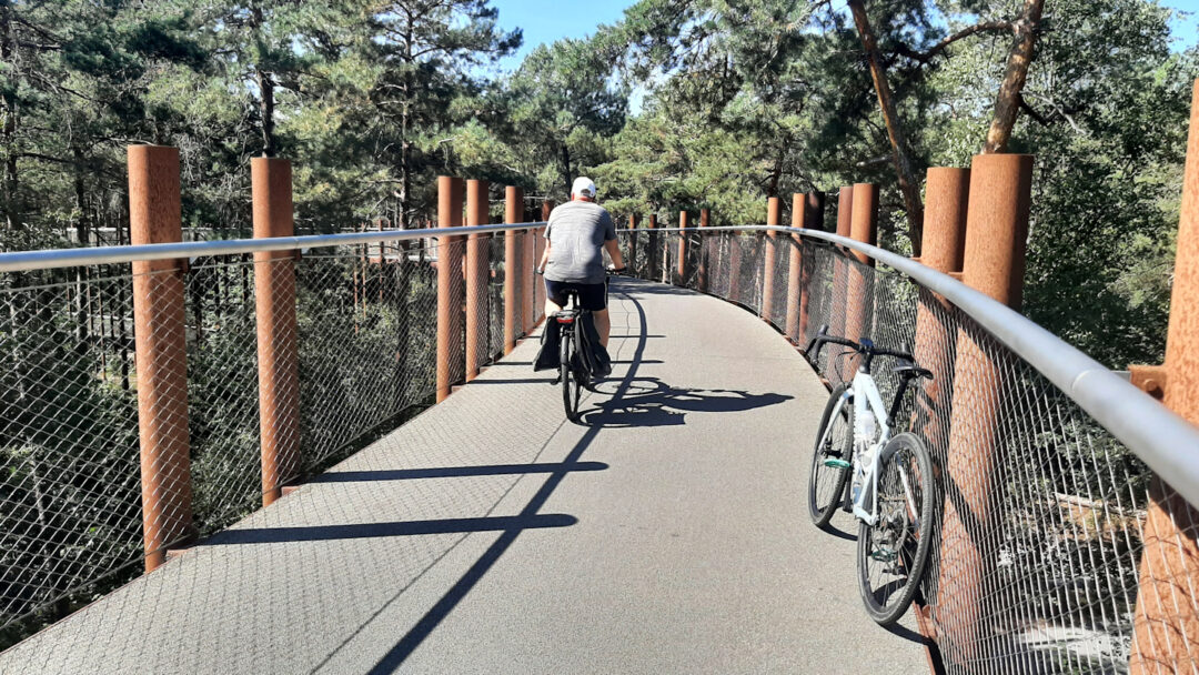 a bicycle leaning on the side fence of a cycle path through the treetops as another cyclist passes by.