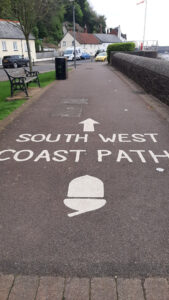 The words "South West Coast Path", and acorn waymark and an arrow pointing ahead, all painted in white on the tarmac footpath