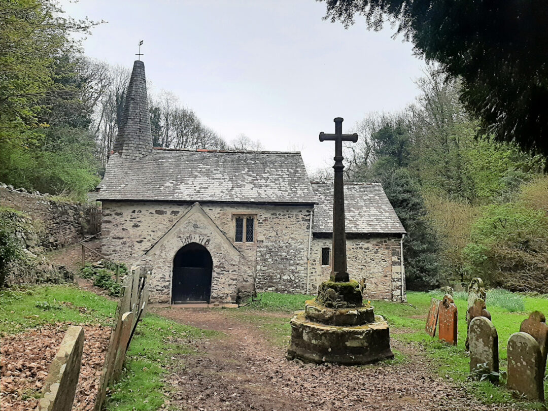 A small church is seen side on (the South side). The church door faces the camera. A stone cross stands in front of the church. The church has a small spire - a 'spirelet'.