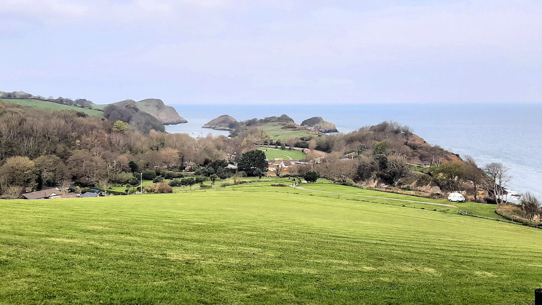 A smooth grassy field slopes down to fields and hedges with a long narrow bay in the distance.