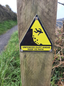 A yellow and black sign with a crude image of a person falling off a stylised cliff, with the words "Unfenced cliff ahead. Keep to Path"