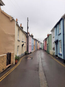 A narrow street with a continuous row of colourful two-storey cottages on either side. It is wet.