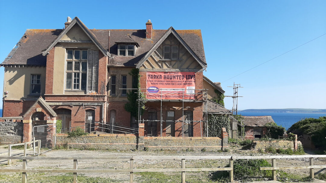 A Victorian Gothic house in red brick with three gables. It is surrounded by security fencing and scaffolding. A large sign advertises a haunting-related charity event.