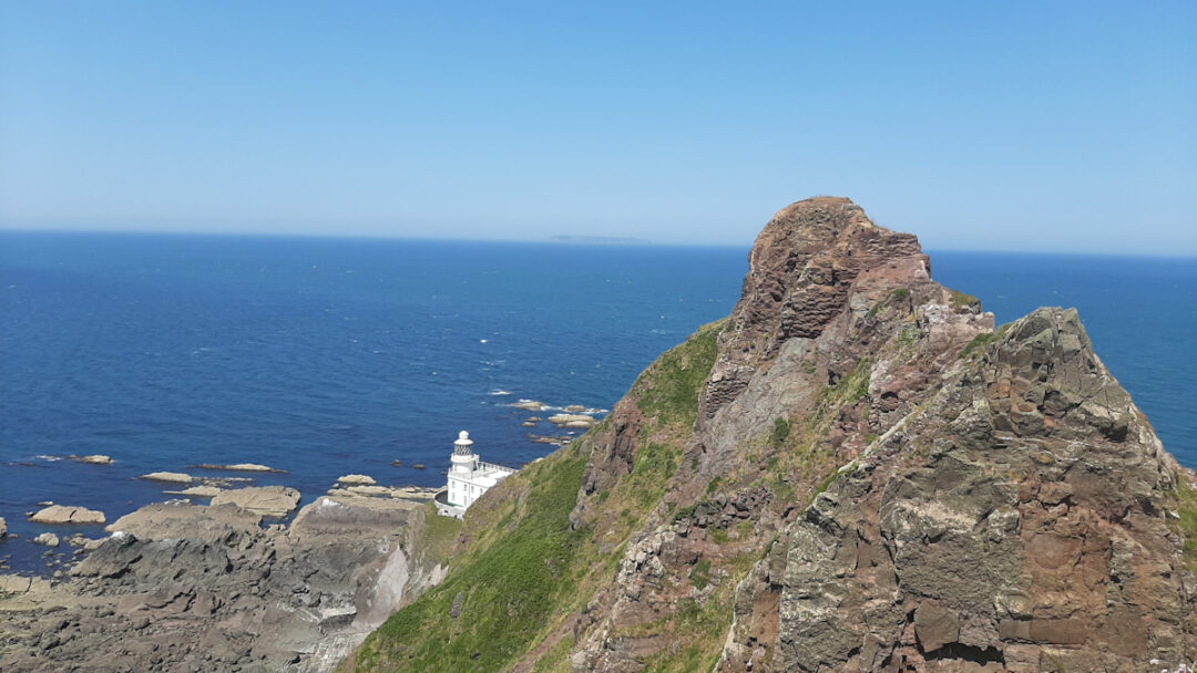 A rocky outcrop in the foreground, a white lighthouse near the water's edge, blue sea beyond.