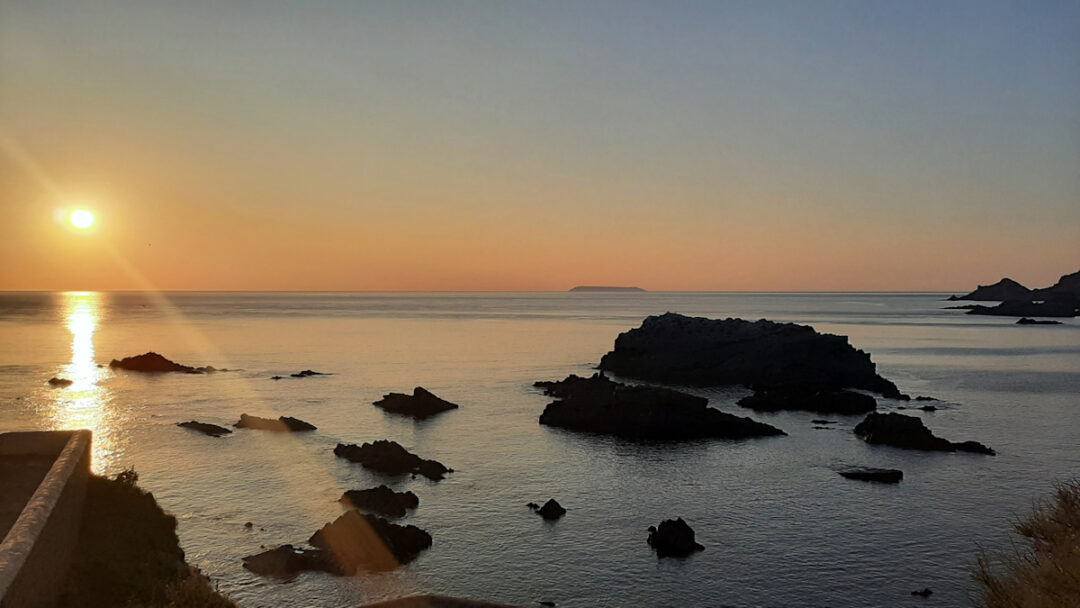 The sun low in the sky over the sea horizon. Orange and purple colours with black rocks silhouetted aginst the sea.