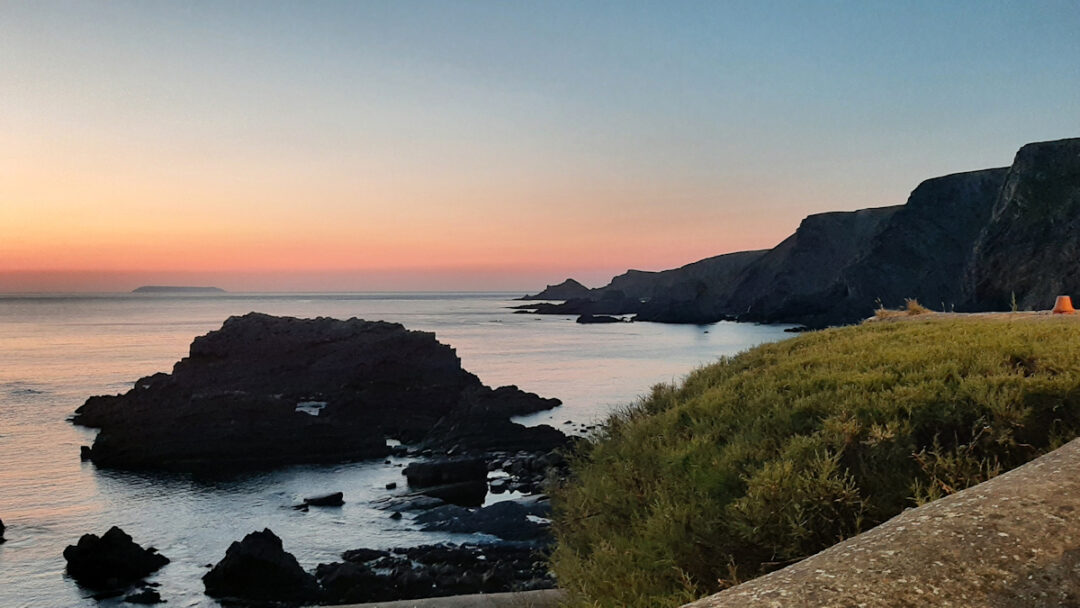 The sky shading from violet to orange over a calm sea. Black rocks and a headland in view.