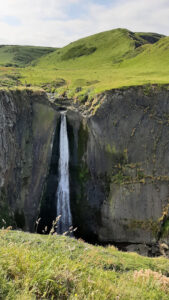 A thin waterfall drops vertically down a dark cliff from green fields