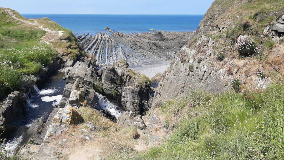 A small stream breaks over stones in a small waterfall. Beyond, straight lines of low rocks stretch out into the blue sea.