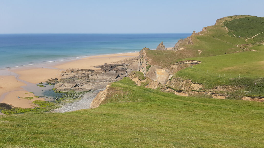 Green heath leads to low cliffs, a sandy beach and blue sea