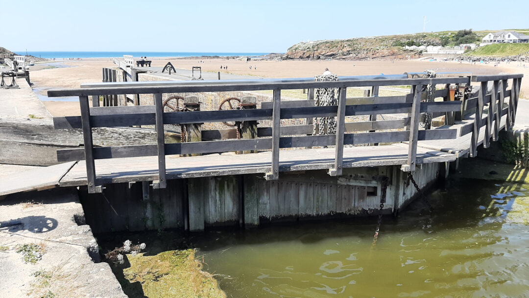 A heavy double lock gate in the near foreground; a wide sandy beach in the distance.