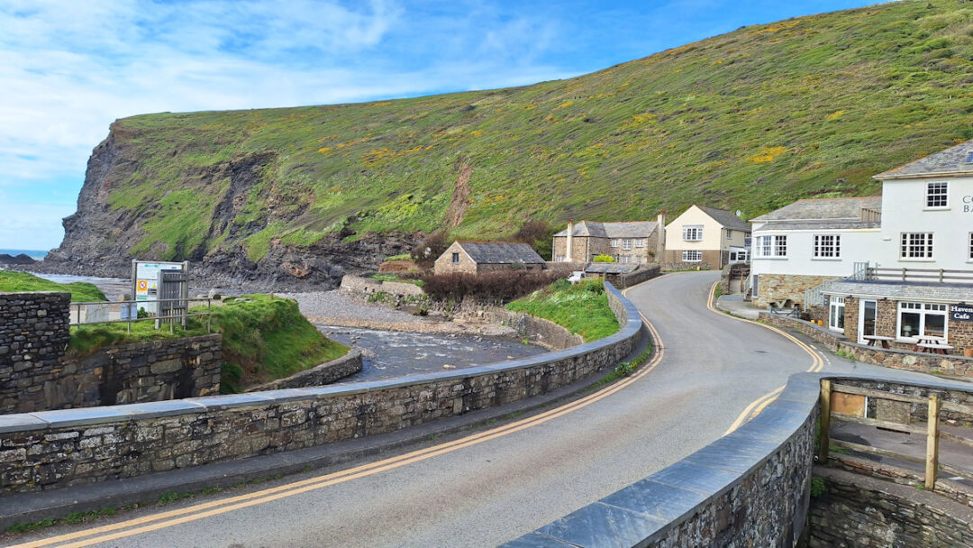 A narrow road curving away, a few buildings, a small stream running to the sea under a low green cliff.