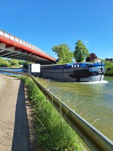 A big blue barge coming towards the camera passes under a road bridge. The sky is a cloudless blue.