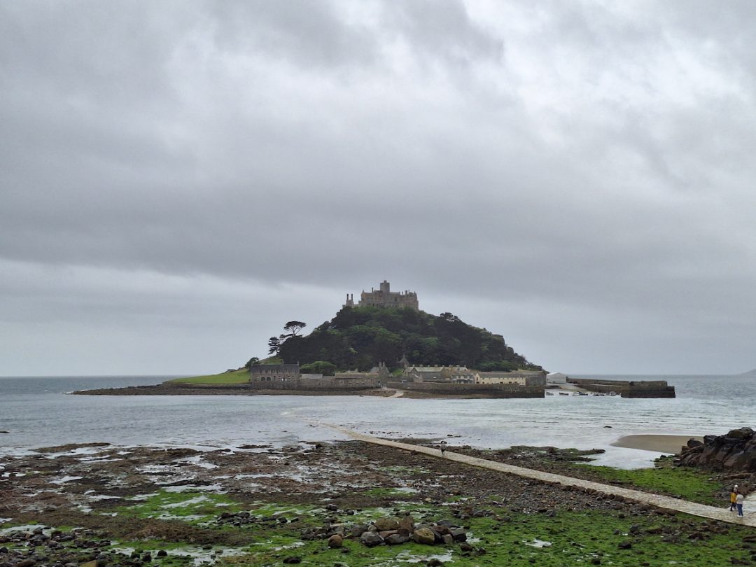 A view of St Michael's mount - a conical-shaped tidal island topped by a castle - seen under a heavy grey sky and set in a grey sea. A stony foreshore in the foreground.