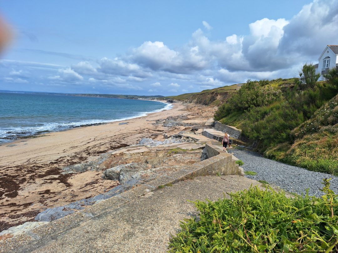 Blue sky, a few white clouds, blue sea, low green cliffs and some concrete ramps leading to a sandy beach