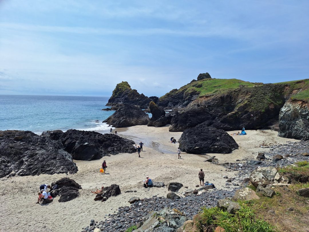A beach. Scattered black rocky outcrops and a green clifftop on the far side. Blue sea and sky. A few people scattered across the beach.