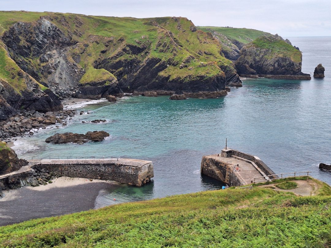 Two stone and concrete jetties form a small harbour with no boats. Calm sea and low green cliffs in the background.