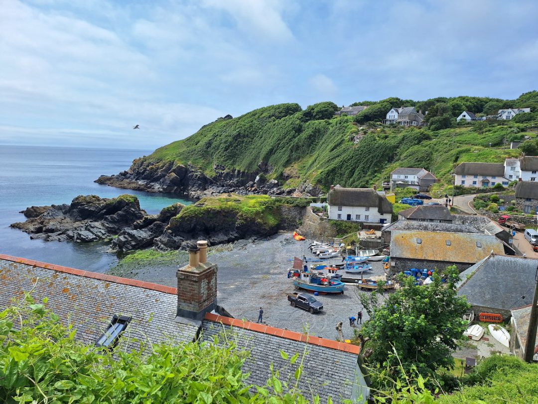 Cadgwith - a small fishing cove with boats hauled up, seen from a point looking over a house roof. Low green cliffs on the far side.