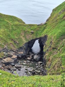 A large stony hole with the sea seen through a rock arch under sloping green cliffs.