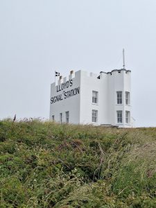 A blocky building painted bright white with ornamental battlements and bearing the words "LLOYD'S SIGNAL STATION" in giant black letters; seen on the skyline on top of a green knoll.
