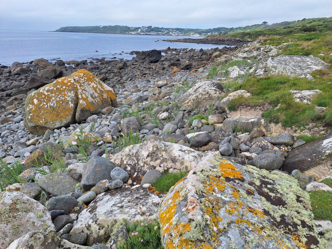 A view along a flat coastline/ Big boulders in the foreground; in the far distance a headland with a village (Coverack)