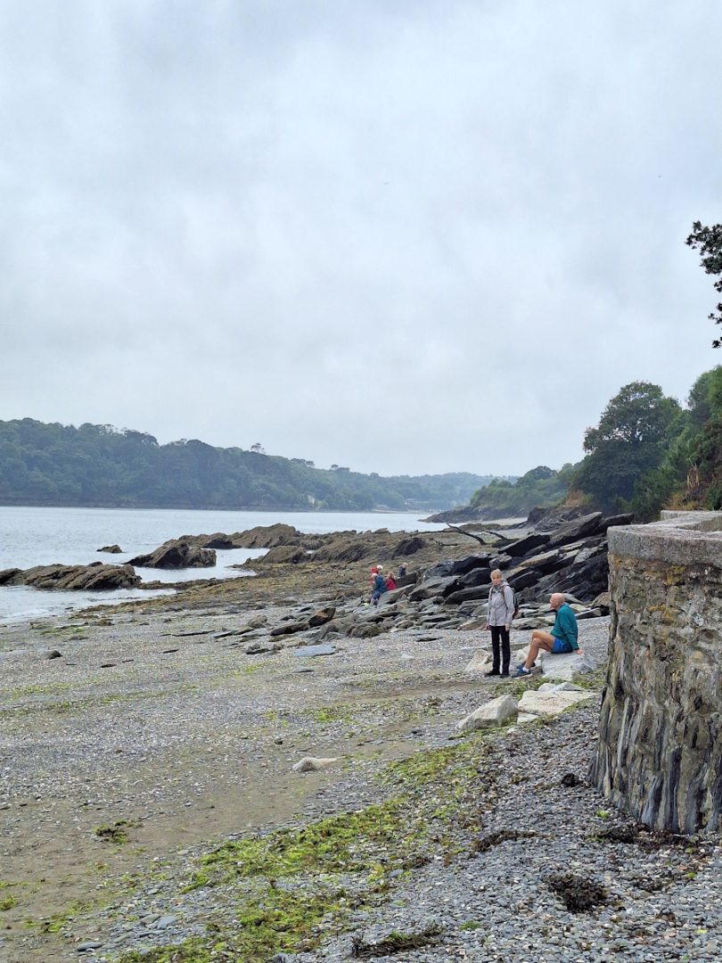 A grey beach - a mixture of pebbles and sand.. A concrete sea wall. Two people. low slabs of rock. Misty distance and light grey sky.