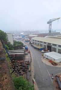 A grey and murky view of commercial docks. Low buildings, a crane, a large cruise ship dominating the scene in the distance.
