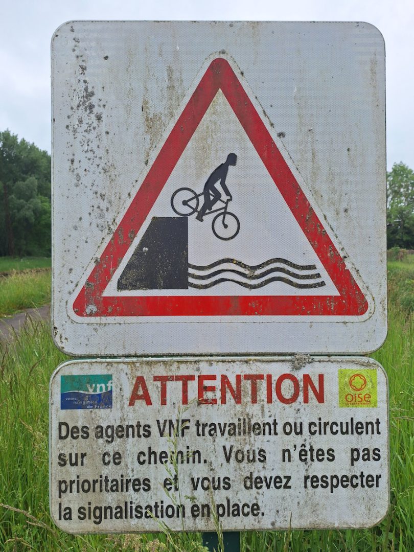 A rather grubby lock-side warning sign showing a cyclist nonchalantly riding off the lock wall into the ricer.