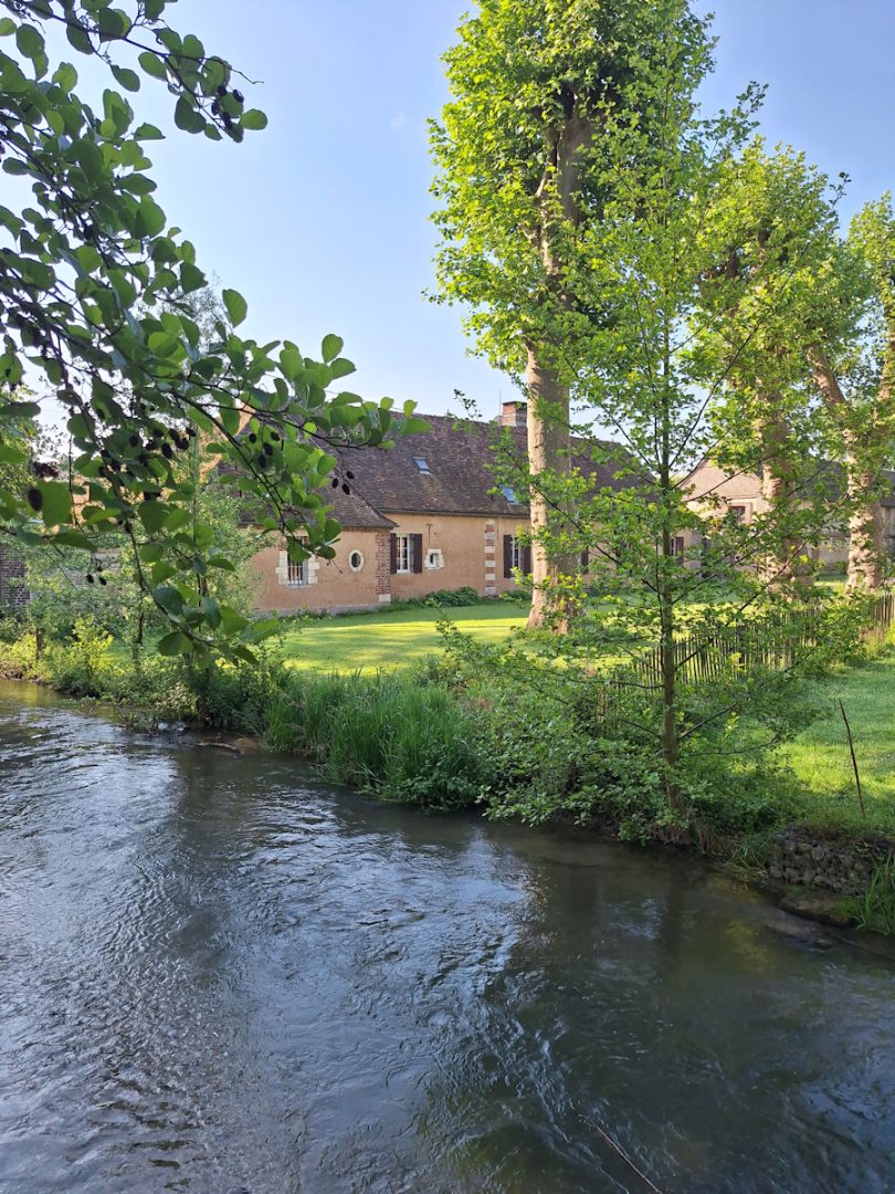 A clear stream in the foreground. On the far bank green trees shade a stone farmhouse with a neat lawn in front