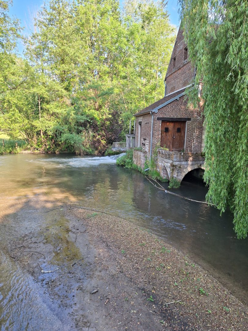 An old stone building on the bank of a clear stream with green tress in the background