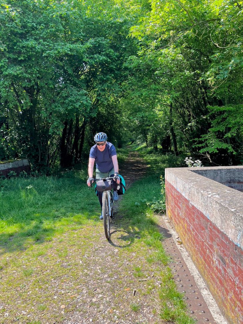 A cyclist in a blue top on a gravel path and emerging from shady woodland. A low wall intrudes on the right of the picture.