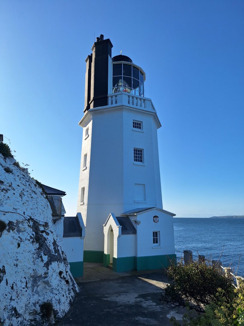 A white octagonal lighthouse. Three sides in shadow, the fourth bright in sunlight. All against a clear blue sky. A rough white cliff on the left of the picture. The lighthouse is crowned with a double chimney behind the light iteself.