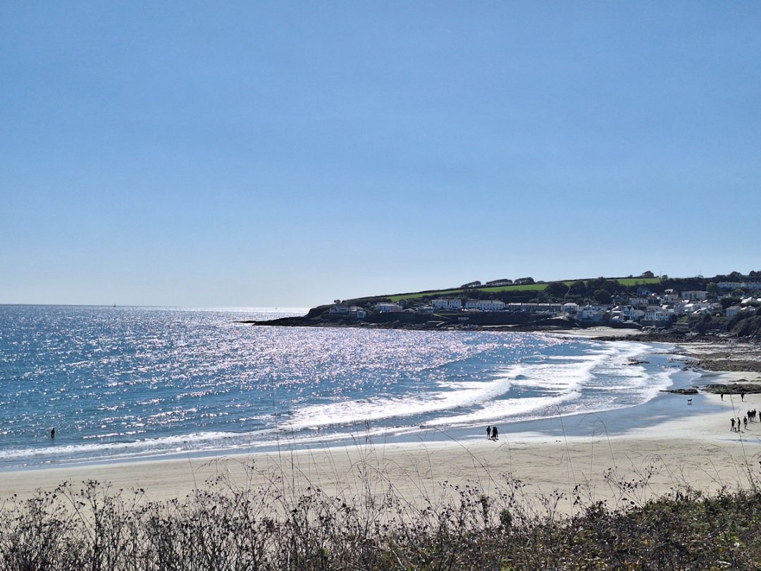 A sandy beach, gentle waves breaking white, a dark headland in the distance, clear blue sky. And the sea bathed in silver light from the sun.