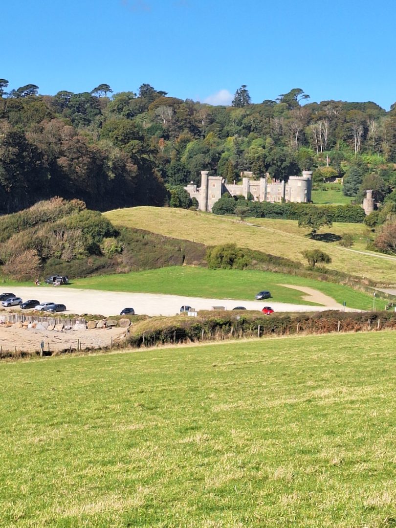 A country house in mock Norman Castle style standing on raised green lawns and fields with a wooded hillside behind. The sun is shining and the sky is blue.