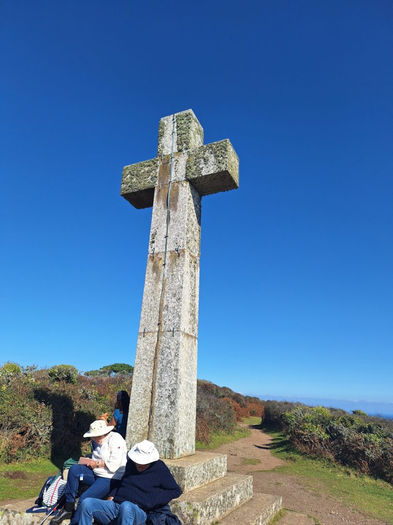 A tall granite cross rears up into a clear blue sky. A couple of random people sit on the steps of the plinth with their faces hidden by their hats