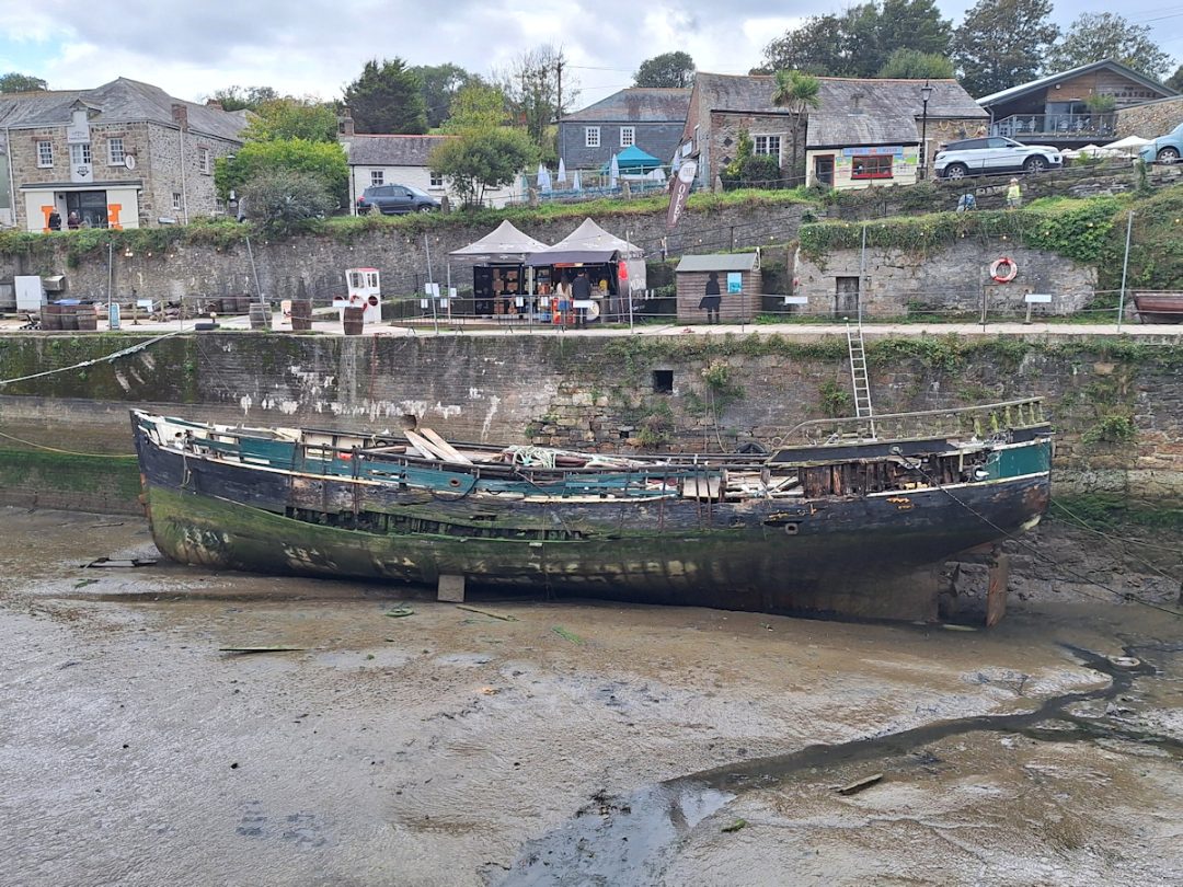 A dock, empty opf water. An old semi-derelict hulk of a ship resting on the mud. Stalls and gazebos on the quayside and a road with various buildings beyond.