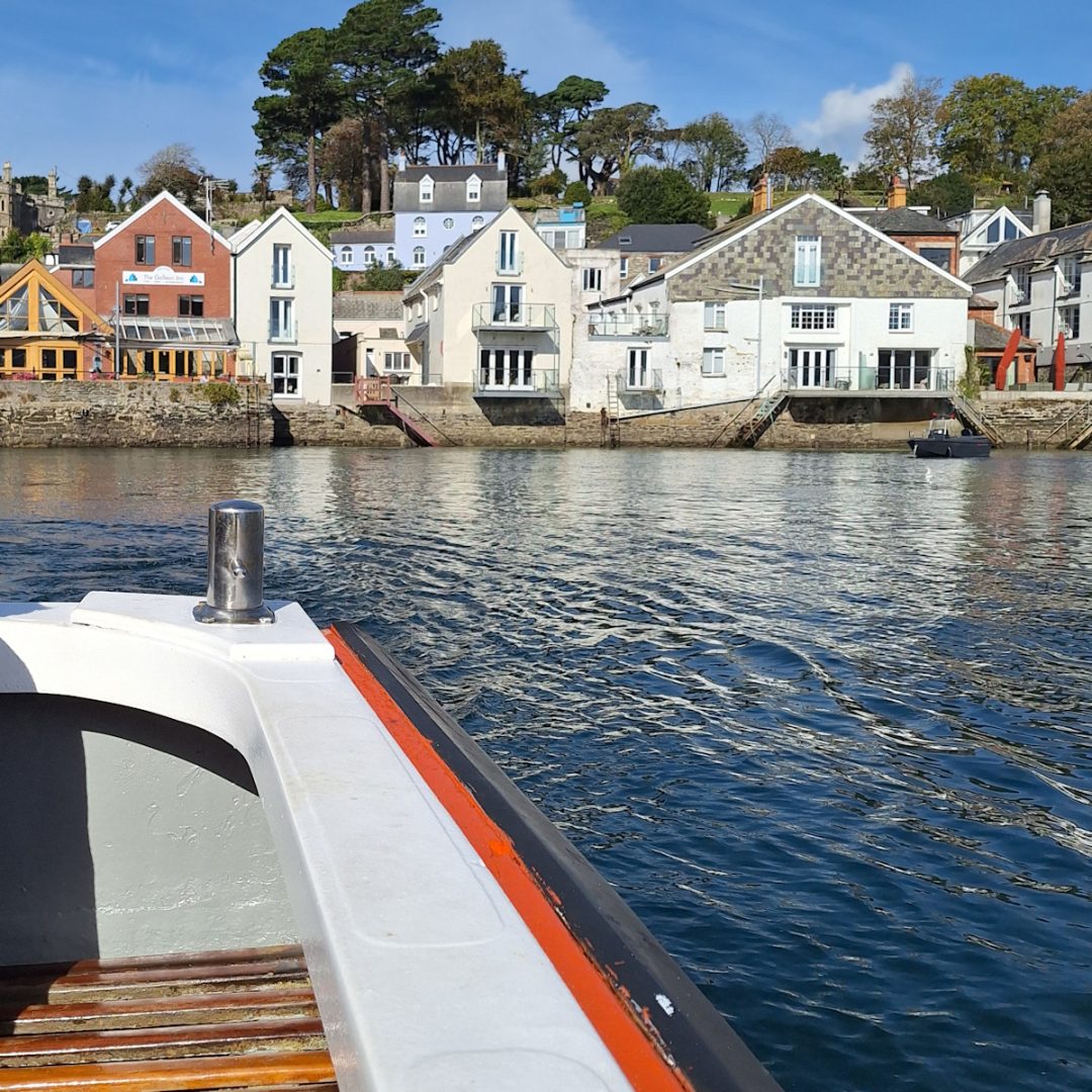 Part of the side and stern of an open boat on calm water, looking back to low buildings on the waterside.
