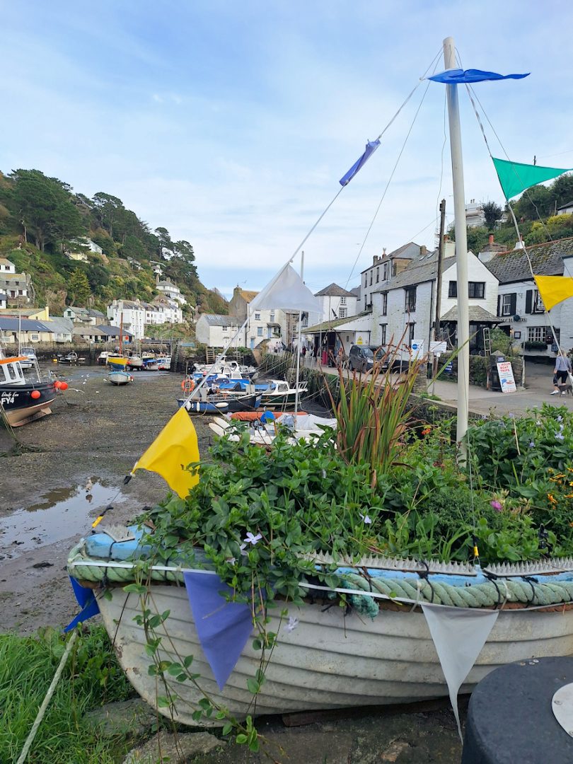 A harbour with several boats, houses and cottages on either side and a boat on dry land in the foreground converted to a planter and decorated with coloured bunting.