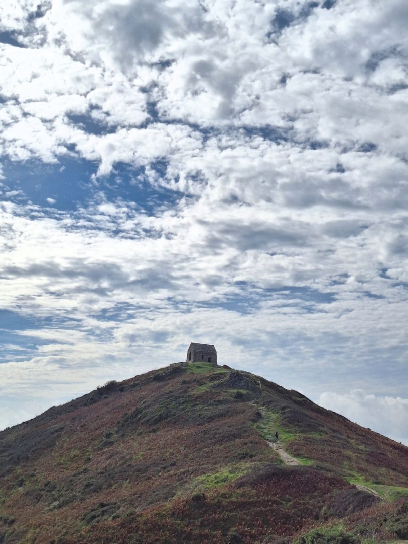 A path winds up a cone-shaped hill covered in low vegetation. A small stone building is silhouetted against a blue sky and silver clouds.