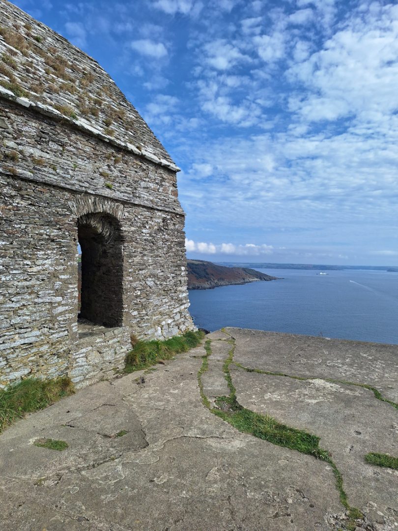Part of an old grey stone building next to a concrete slab, with a view along the coast.