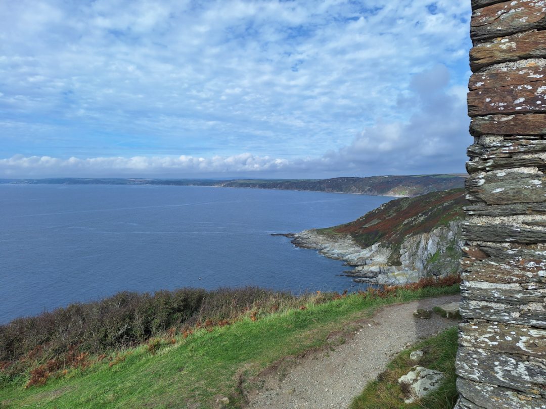 A coastline with low green cliffs disappearing into the distance. Calm blue sea on the left, blue sky with thin white clouds above.