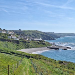 A green path between low wire fences leads along a low green cliff to a small village. The sky is blue with a few wispy white clouds. Below the village a beach of grey sand leads to a small harbour with two low curved enclosing walls of grey stone. The tide is out and the harbour is just sand; there are no boats. The sea on the right is calm with a few white waves breaking. The low cliffs continue along the coast into the distance.