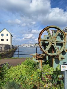 in the foreground, and old iron winding gear with cogs. Further away, the end of a modern white apartment block. In the far distance across the water a large cruise ship, all under a blue sky part-covered in white clouds.