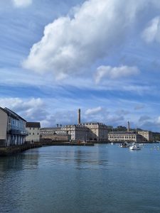 Centred in the picture a group of solid-looking grey stone buildings fills the frame left to right. The water below is calm and blue. The sky is also blue with white clouds.