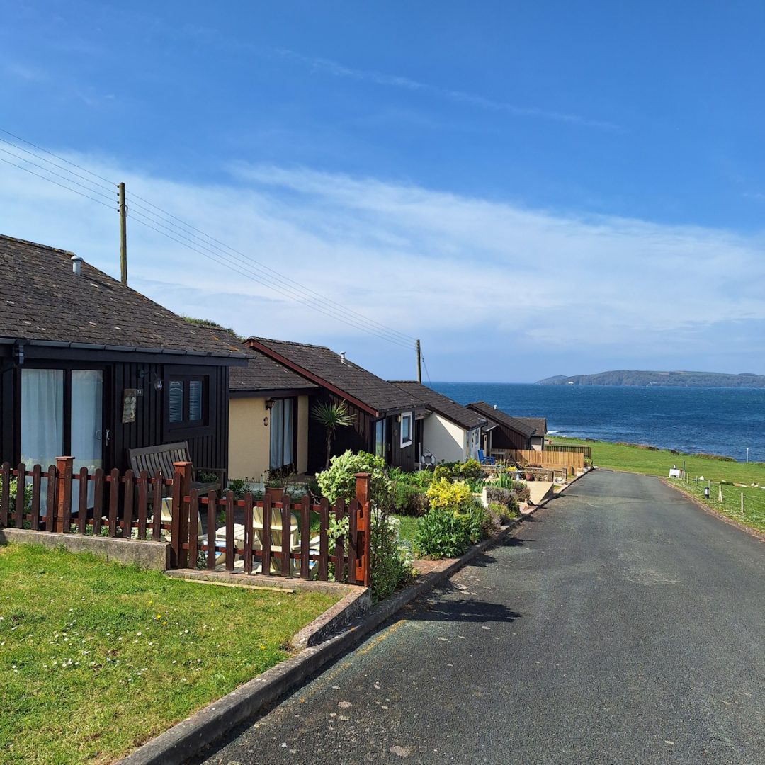 Looking down and along a row of single storey holiday bungalows, each with a small well-kept front garden of grass or flower beds. A tarmac road runs in front. In the distance acroos a bay of blue sea a low headland can be seen under a blue sky with some white cloud.