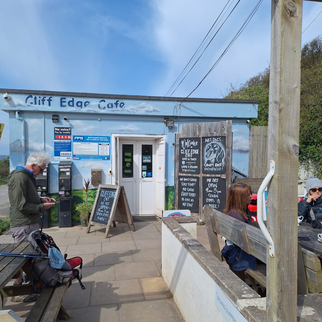 A small, pale blue flat-roofed single-storey shack with "Cliff Edge Cafe" (no accent) along the roof fascia. Narrow white entrance door. Paved area outside with tables and a few customers. Chalked notices by the door include one with a portarait of a dachshund saying "Wet Dogs Sandy Boots Encouraged".