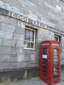 The wall of a grey stone building fills the frame. Two sash windows, one mostly obscured by a standard red telephone box. Above the windows the word 'COOPERAGE' has been painted and then overpainted in a darker colour and more closely spaced so the word shows twice.