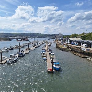 Three lines of pleasure craft moored at jetties in the river. Low buildings on the right. Calm water, blue sky with puffy white clouds.