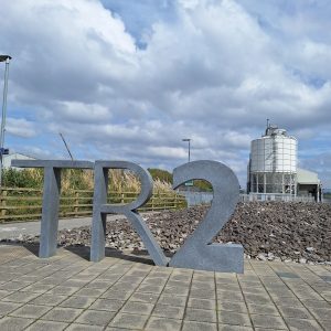 Three stones about a metre and a half high spell "T R 2". Beyond, a "beach" of large grey pebbles. To the left a wooden fence and tall grasses, and further away a white metal tower. Blue sky with many white/grey clouds.