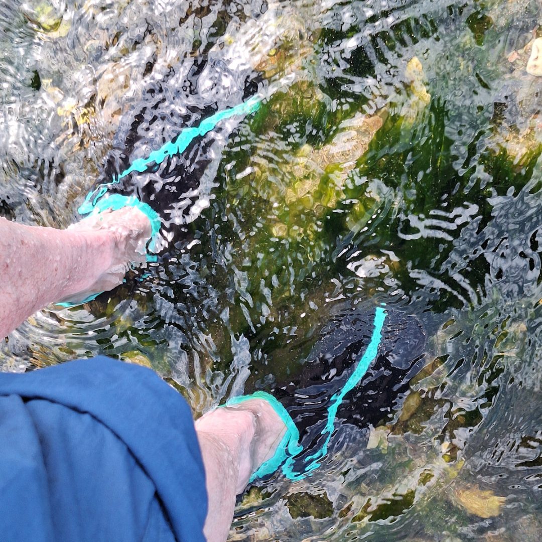 A pair of white legs, one with a rolled-up trouser leg, standing in clear running water which doesn't reach the knees. On the feet are strange black shoes with a pale blue stripe.
